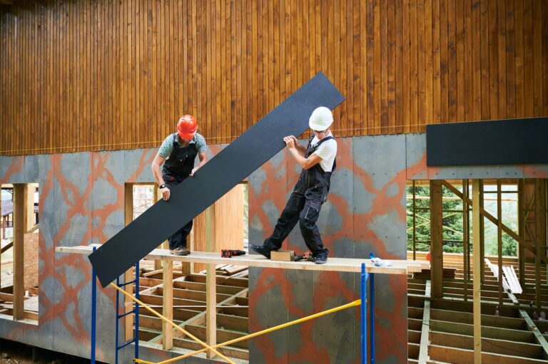 Workers cladding facade of house with cement particle boards while constructing wooden frame house.